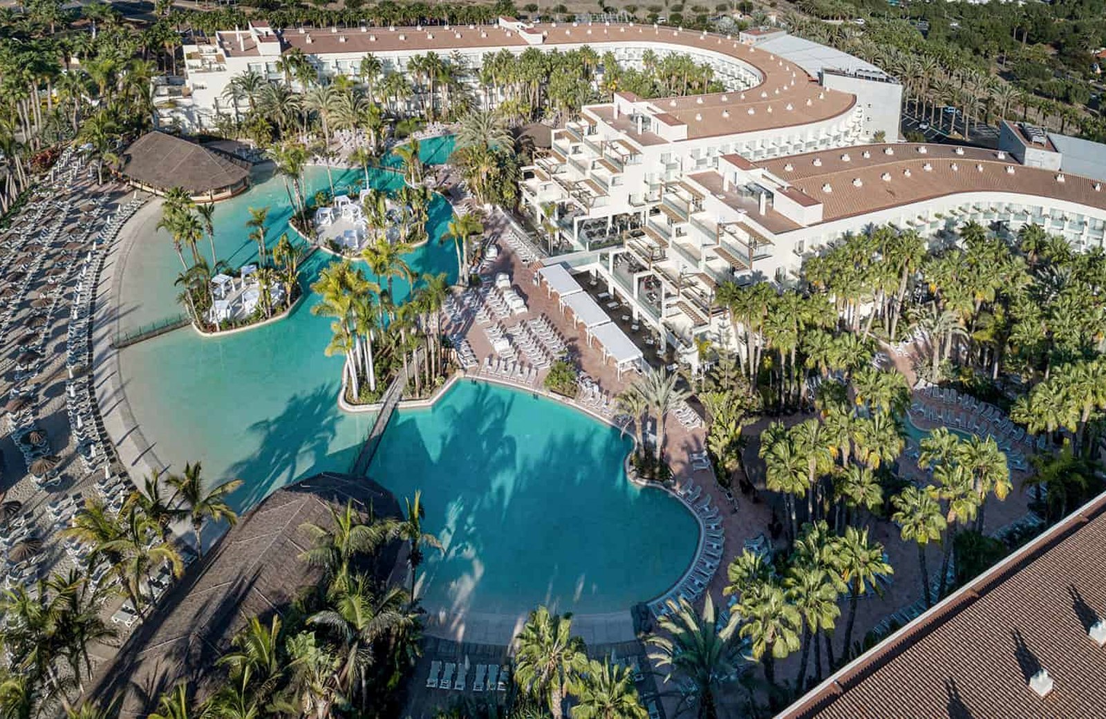 Tabaiba Princess from the air: low white hotel blocks wrapped around a lagoon-shaped pool complex planted with tall palms, with a sandy artificial beach and rows of sun loungers along one side.