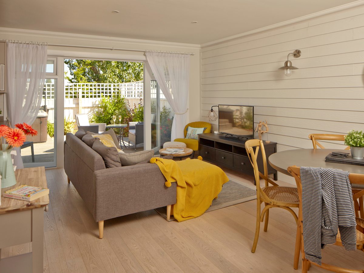 Open-plan living room with a grey sofa and yellow throw, a television on a dark sideboard, a round dining table with cane chairs and sliding doors open to a furnished patio.