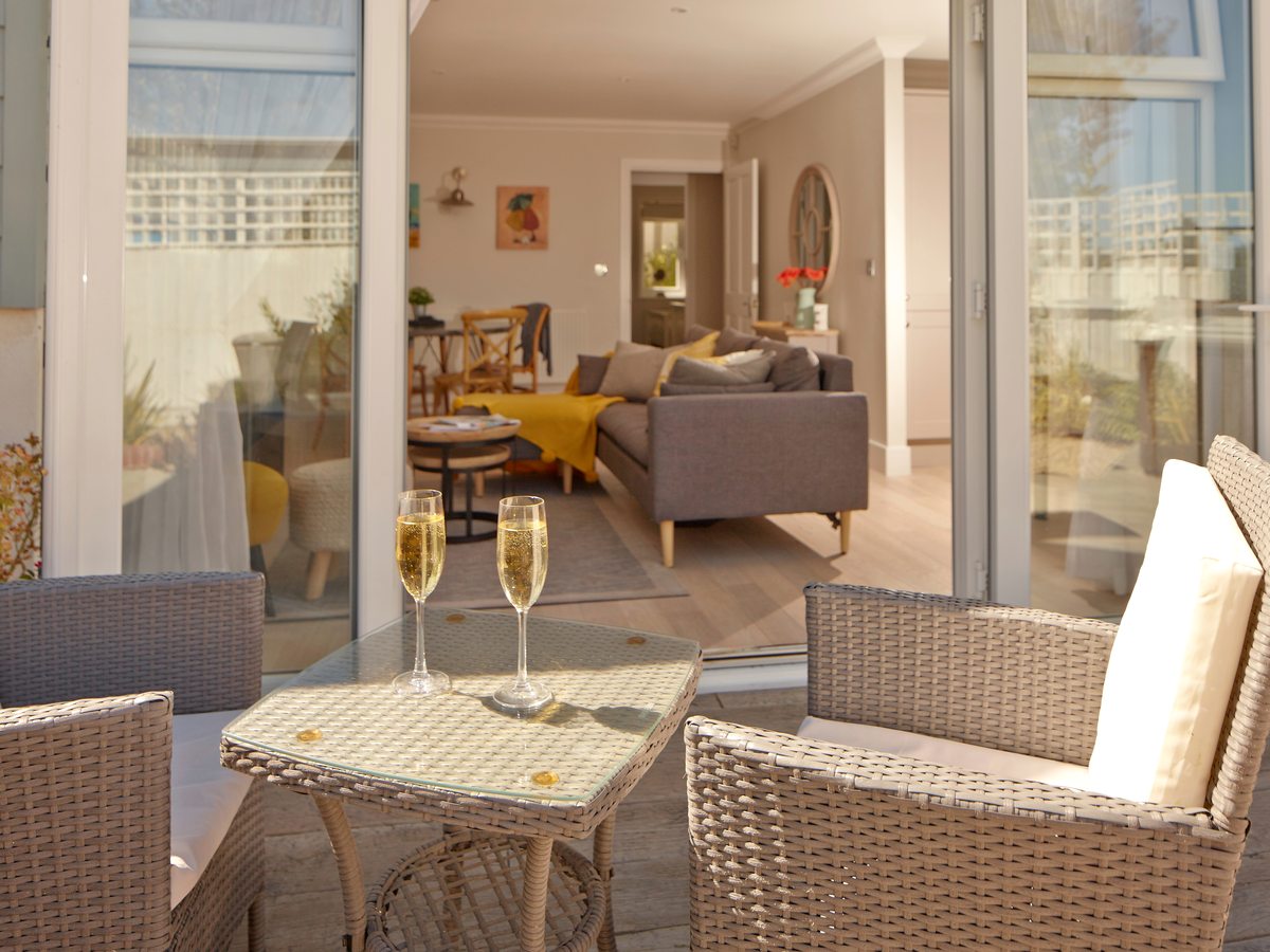 Rattan armchairs and a glass-topped table on a paved terrace, with sliding doors open into the living room behind.