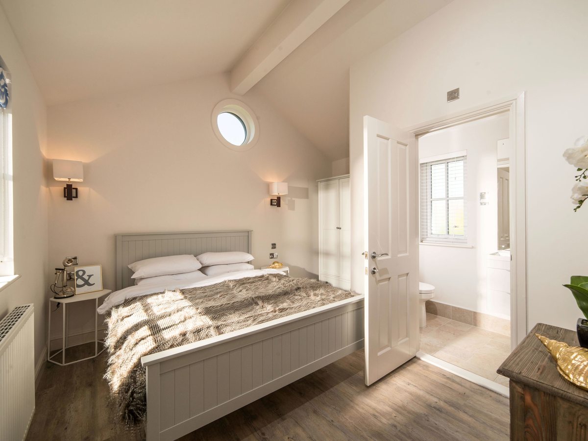 Bedroom with a grey wooden double bed under a vaulted ceiling with a round porthole window, a wardrobe alongside and an open door through to a bathroom.