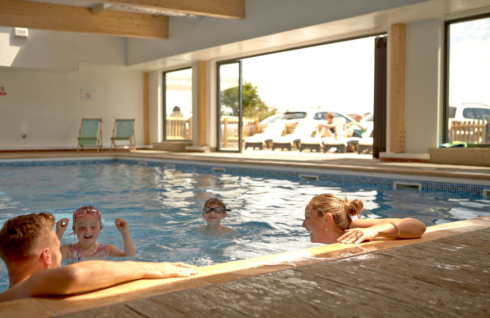 Two adults and two children swimming in the indoor heated pool at The Bay Colwell, with glass doors folded back onto a decked terrace of sun loungers.