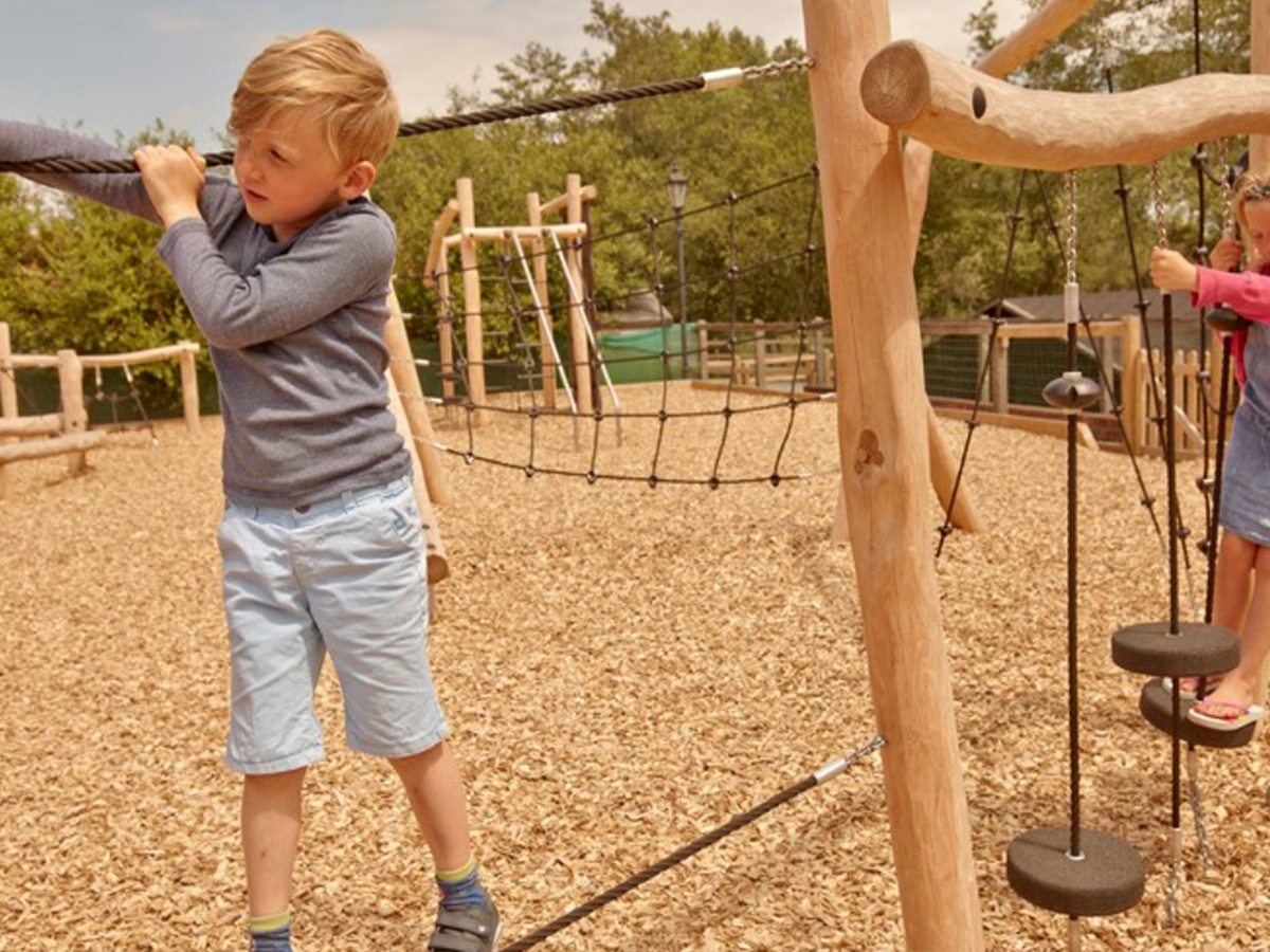 Children climbing on a timber adventure playground with rope bridges and hanging stepping discs over a woodchip surface.