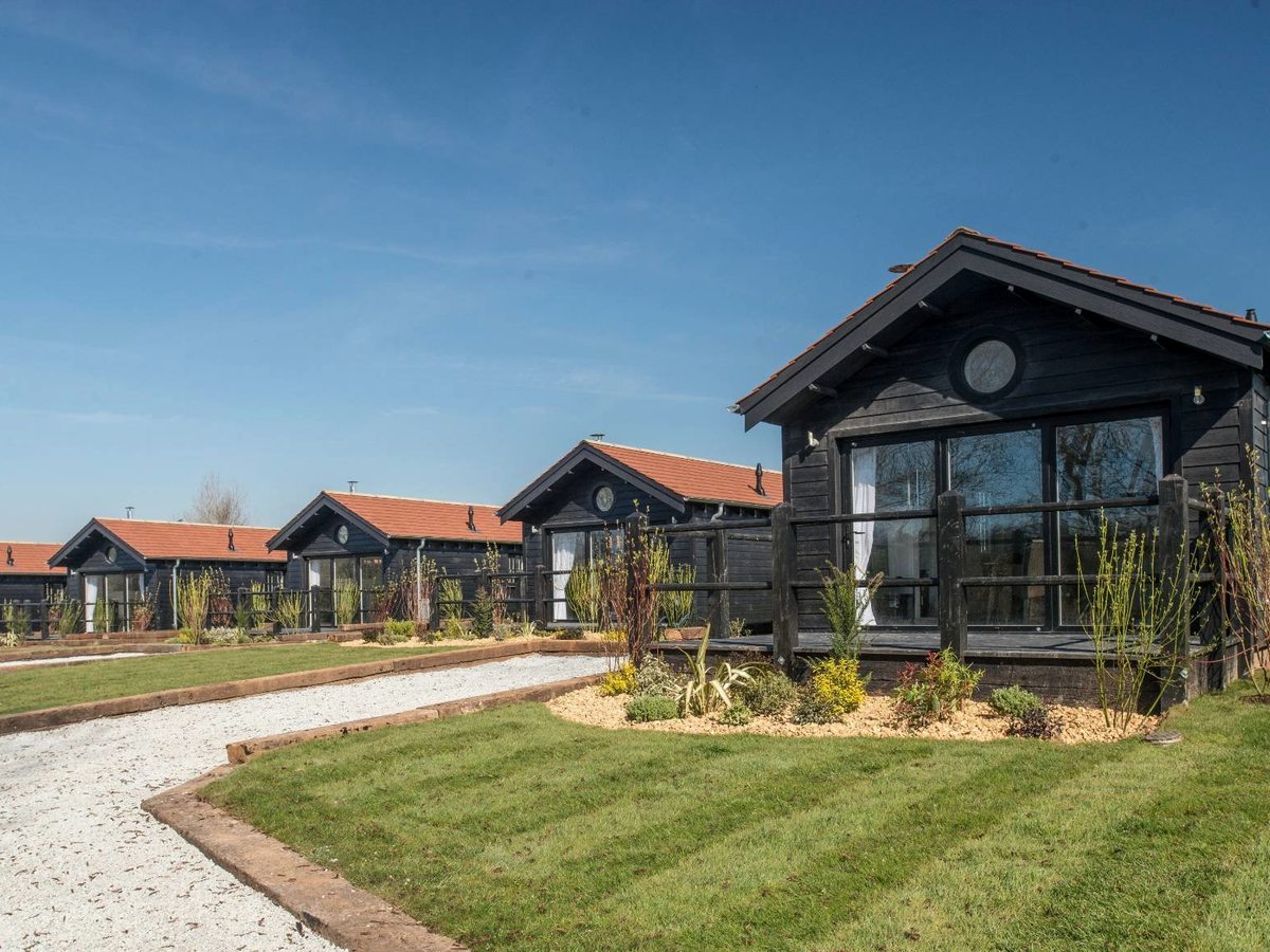 A row of detached black timber lodges with red pantile roofs, full-height glazed gables and decking, along a gravel path with mown lawns.