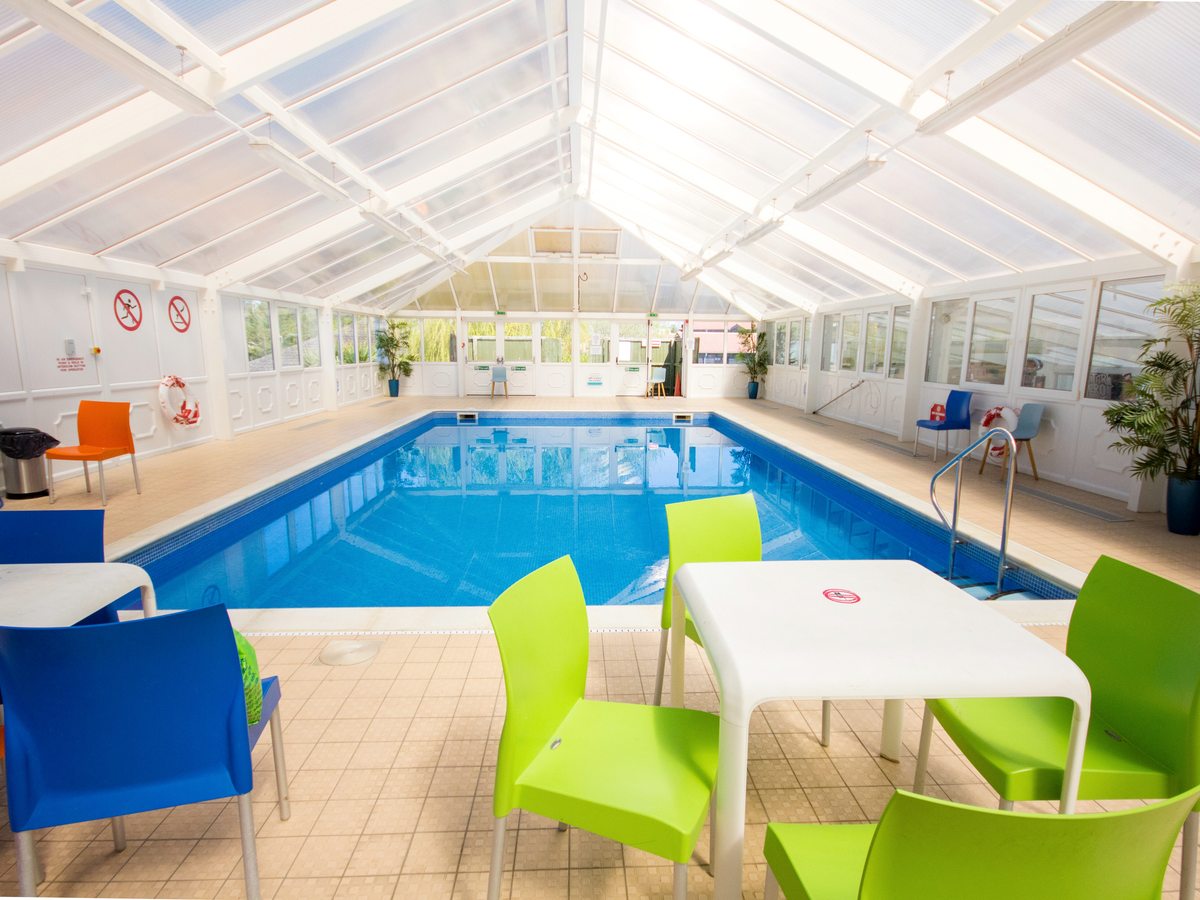 A rectangular indoor pool under a white pitched glazed roof, with a tiled surround and tables with green, blue and orange plastic chairs.