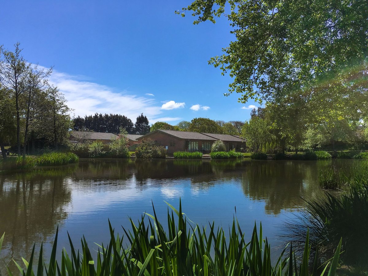 A calm lake fringed with reeds and trees, with a low brick building on the far bank under a blue sky.