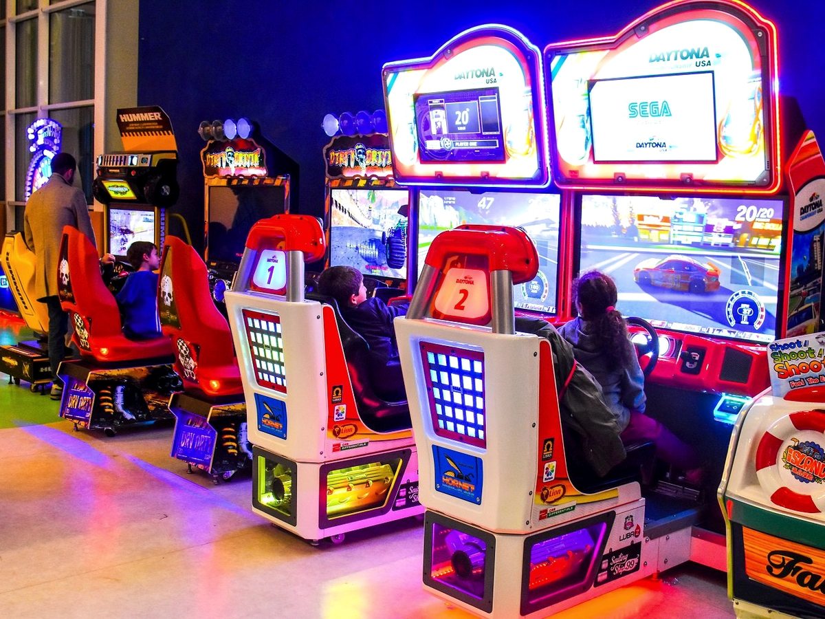 Children sitting in racing-game seats at a row of brightly lit arcade machines.