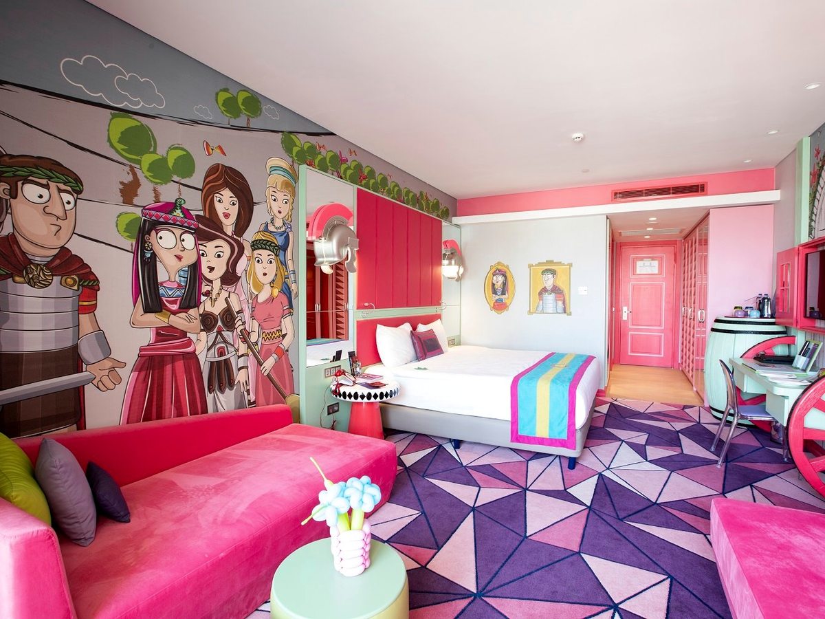 A hotel room in the pink theme with a double bed, a coral headboard panel, a cartoon mural of characters across one wall and a pink sofa.
