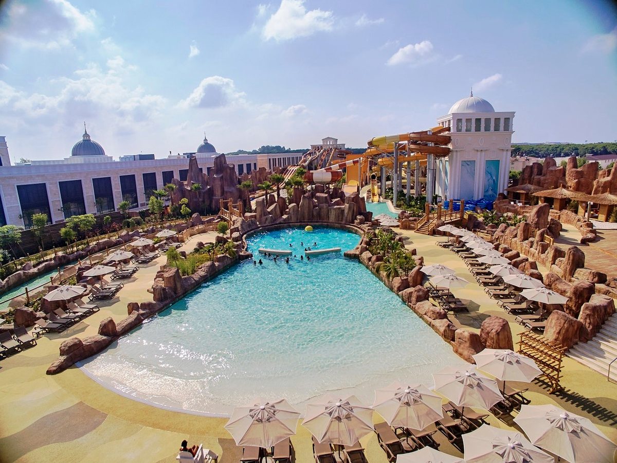 A large outdoor wave pool with a sloping sandy entry, edged with artificial rockwork, ranks of sun loungers and parasols, and water slides behind.