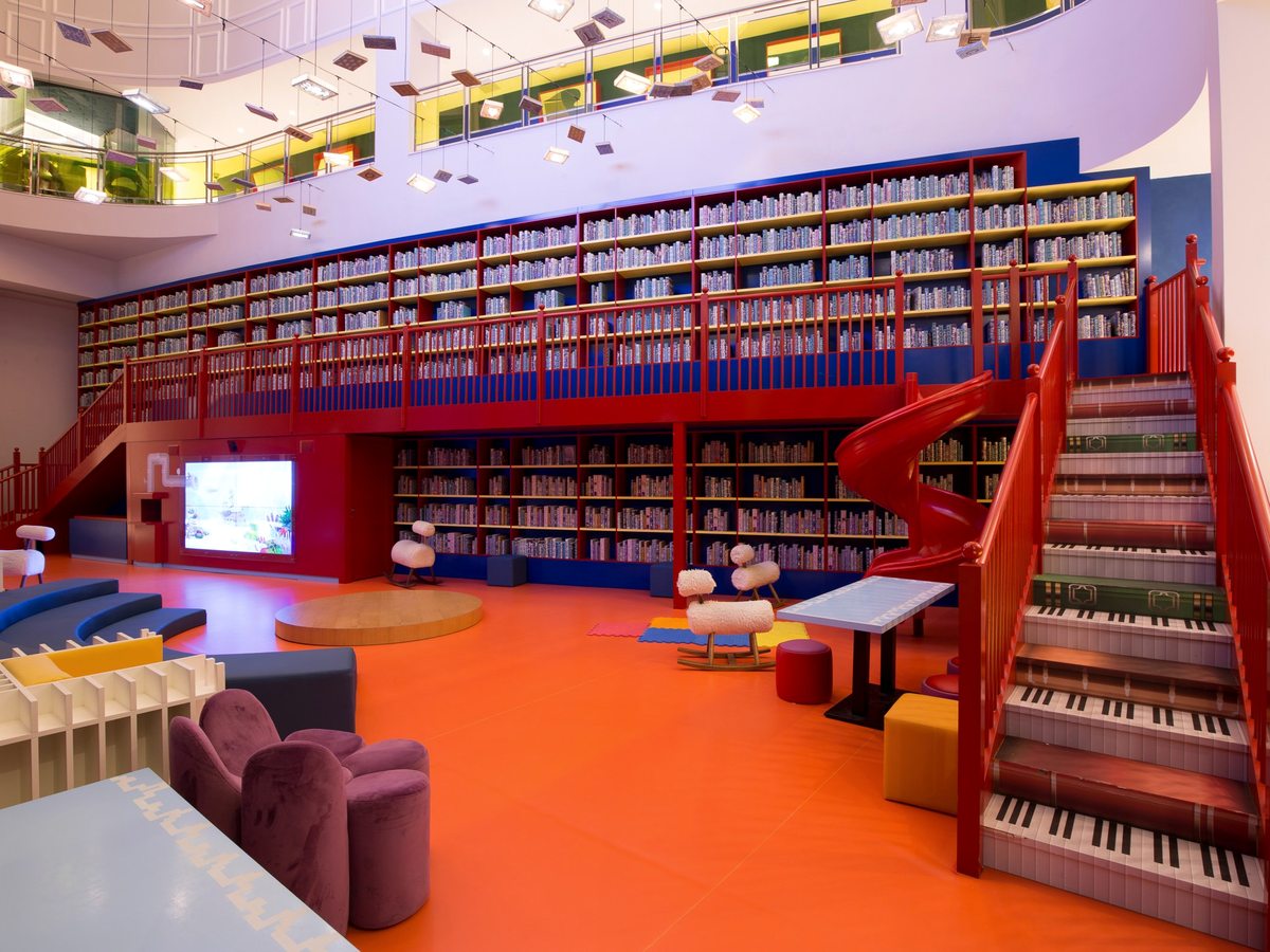 A double-height children's club with a wall of bookshelves, a red spiral slide, a piano-keyboard staircase and low seating on an orange floor.