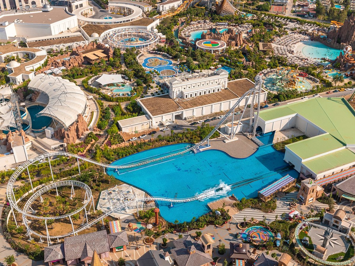 An aerial view of the theme park, with a roller coaster looping beside a large blue lake, the dolphinarium tents to the left and the water park's pools, lazy river and slide towers behind.