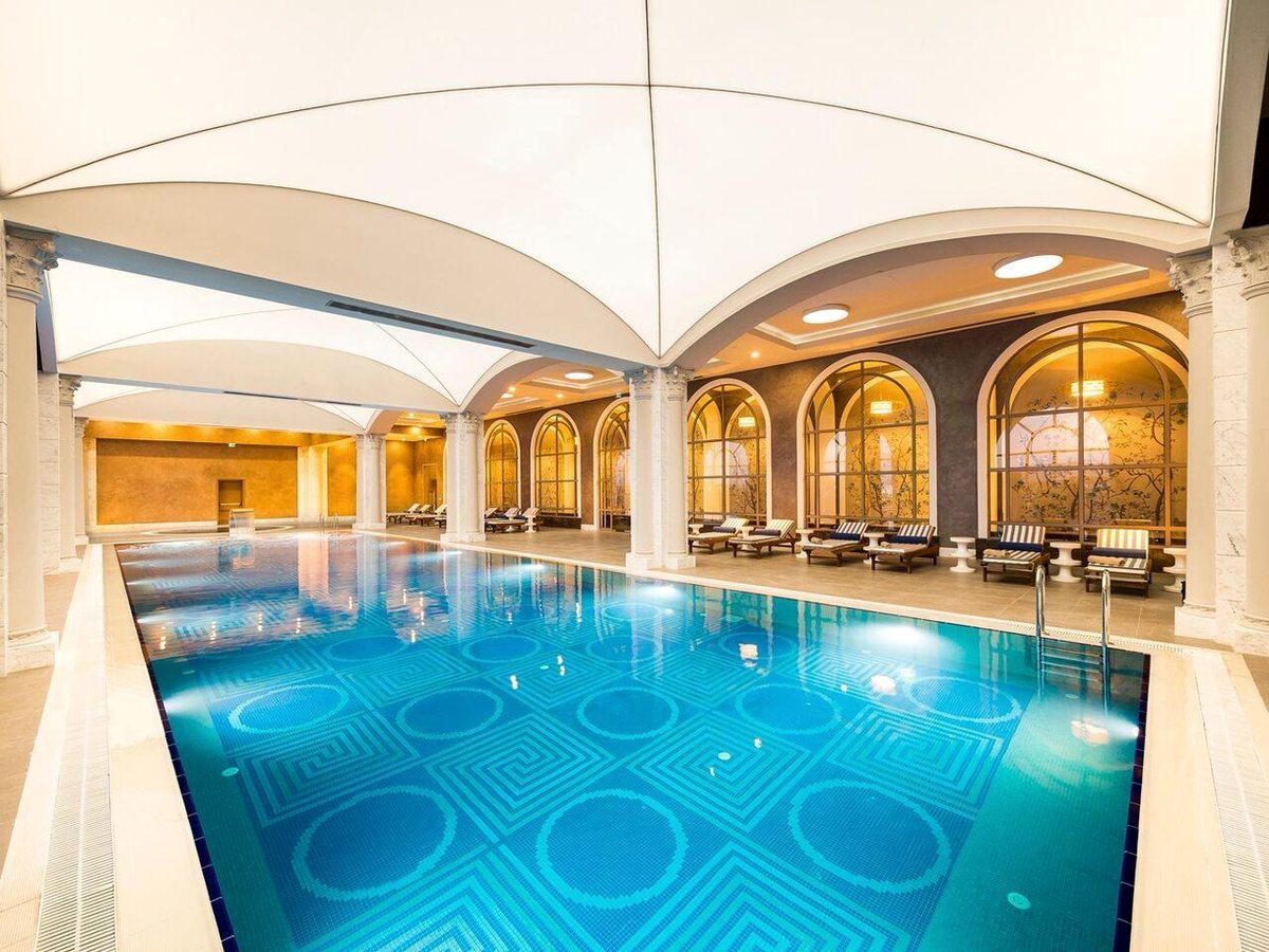 A long indoor pool with a patterned blue mosaic floor under a white vaulted ceiling, loungers along a tiled deck beside tall arched windows.