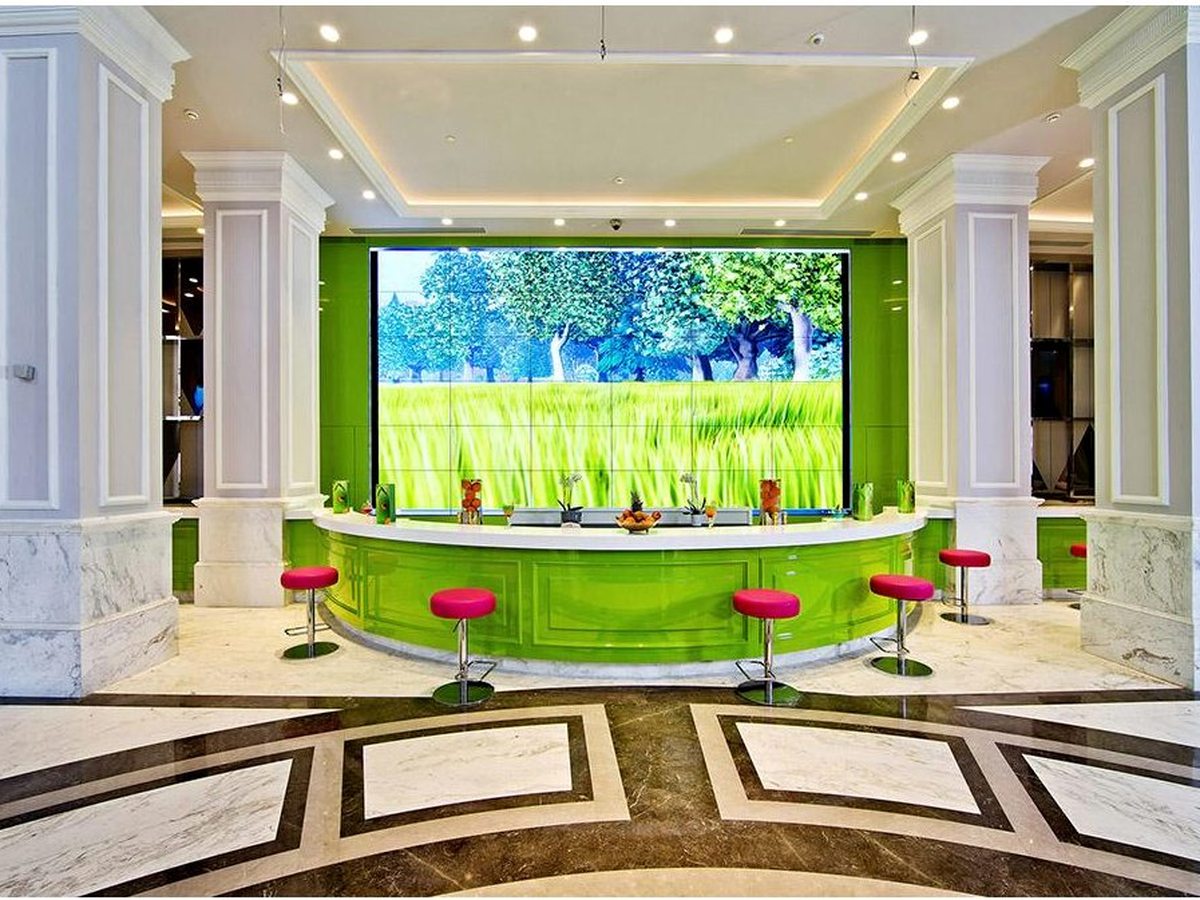 A curved lime-green bar counter with pink stools on a marble floor, a large video wall showing a meadow scene behind it.