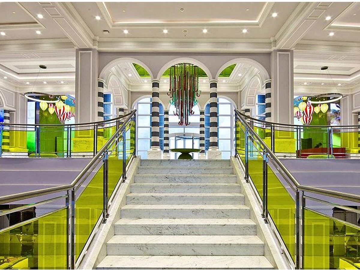 A wide marble staircase between yellow glass balustrades, rising to an arched atrium hung with a red and green chandelier.