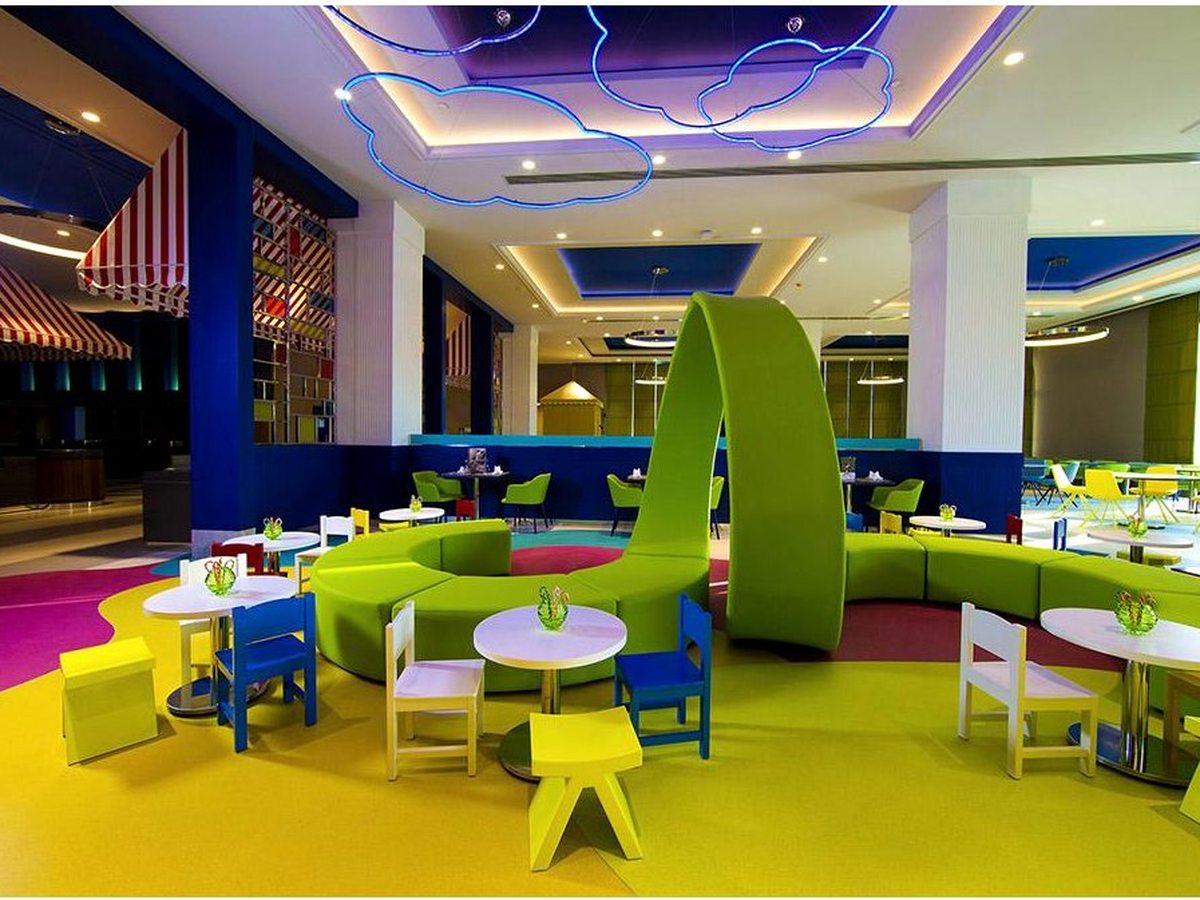 A children's dining area with low white tables, small blue and yellow chairs and a curved lime-green banquette, under blue neon cloud lights.