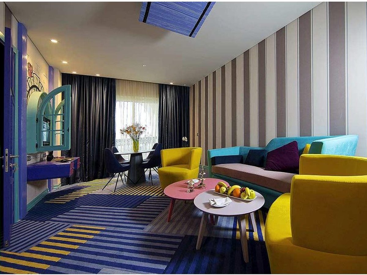 A suite living room with a striped teal sofa, yellow armchairs, low round tables and a dining table for four by the window.