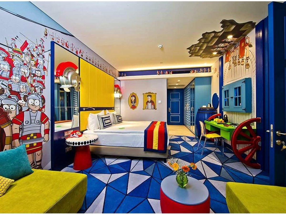 A hotel room in the blue theme with a double bed, a yellow headboard panel, cartoon soldier murals, a blue geometric carpet and a lime sofa.