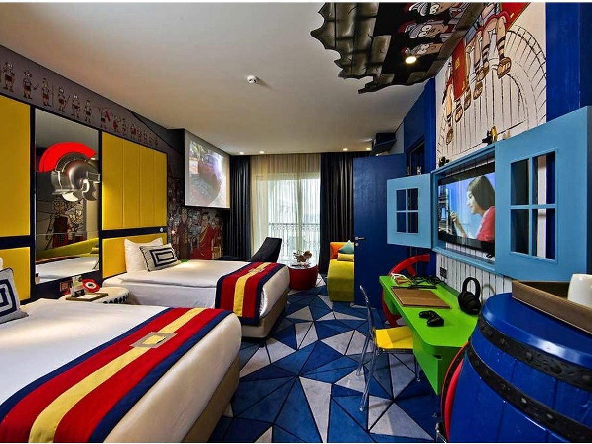 A hotel room in the blue theme with two single beds under striped throws, a green desk with a games controller and a projector screen above.