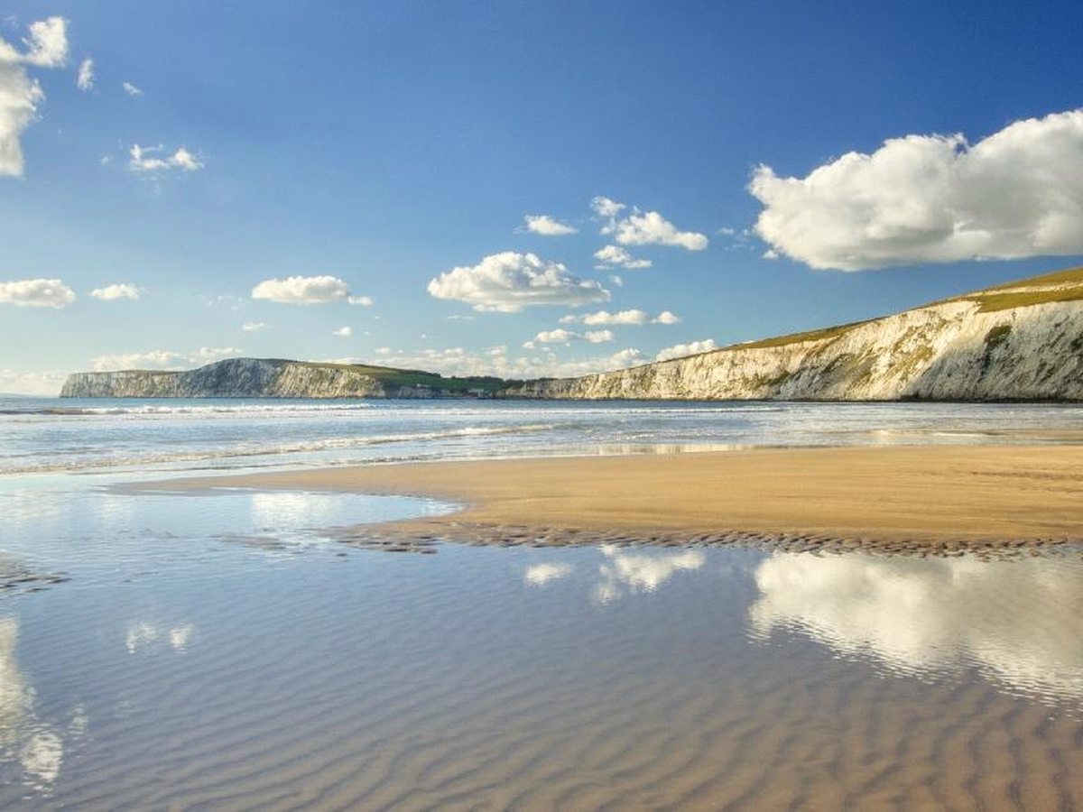 A wide sandy beach at low tide with clouds reflected in the wet sand and white chalk cliffs curving away along the bay.