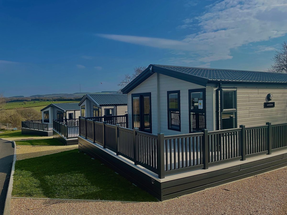 Two modern grey and cream holiday homes with raised timber decking on a mown lawn under a blue sky.
