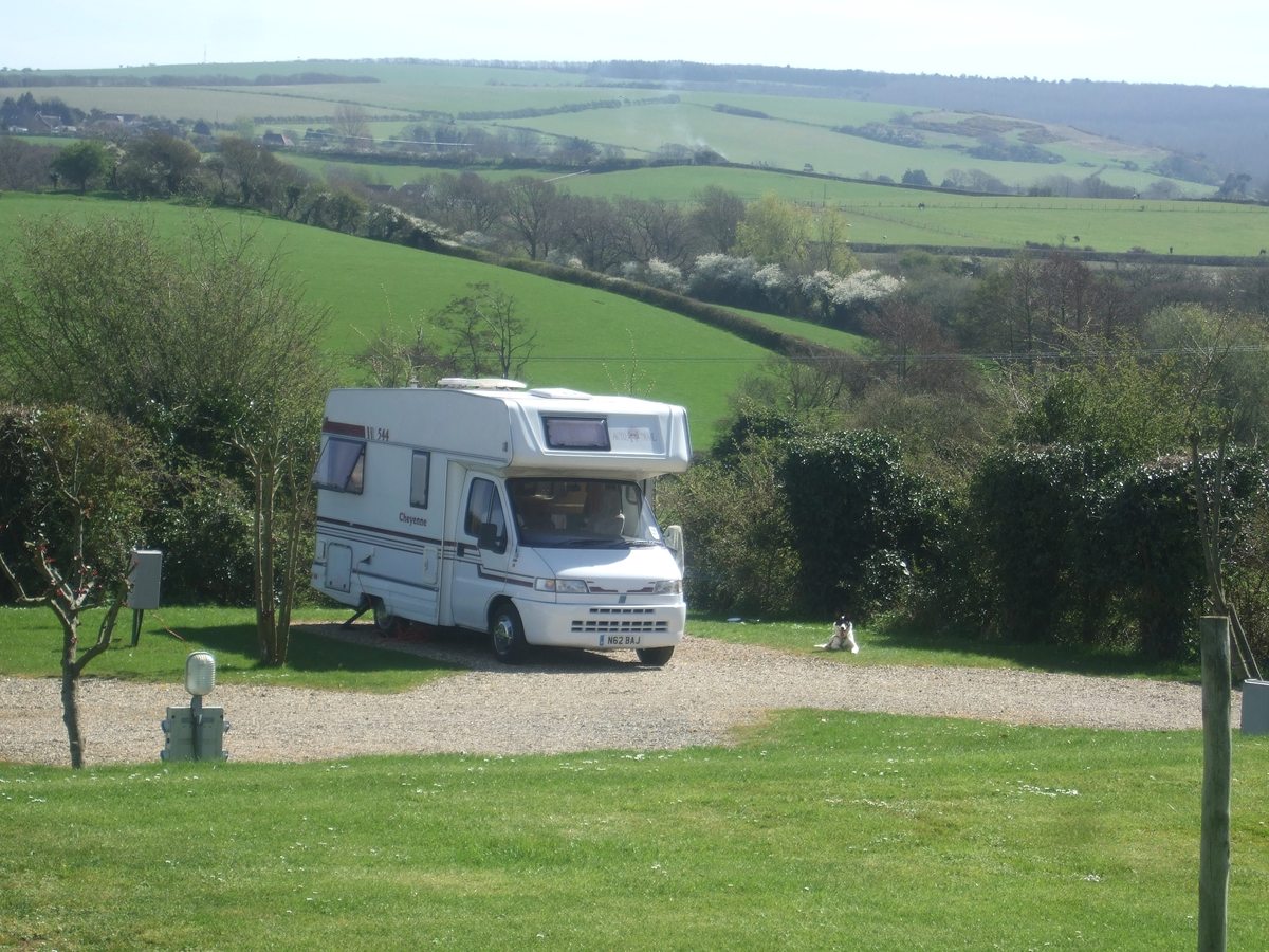 A white motorhome on a gravel hardstanding pitch beside a hedge, with green farmland rising behind.