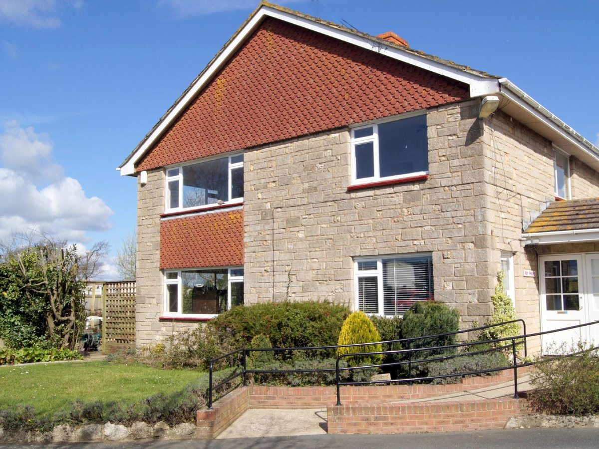 A two-storey stone house with a red-tiled roof and white windows, with a metal handrail beside a ramped path to the door.