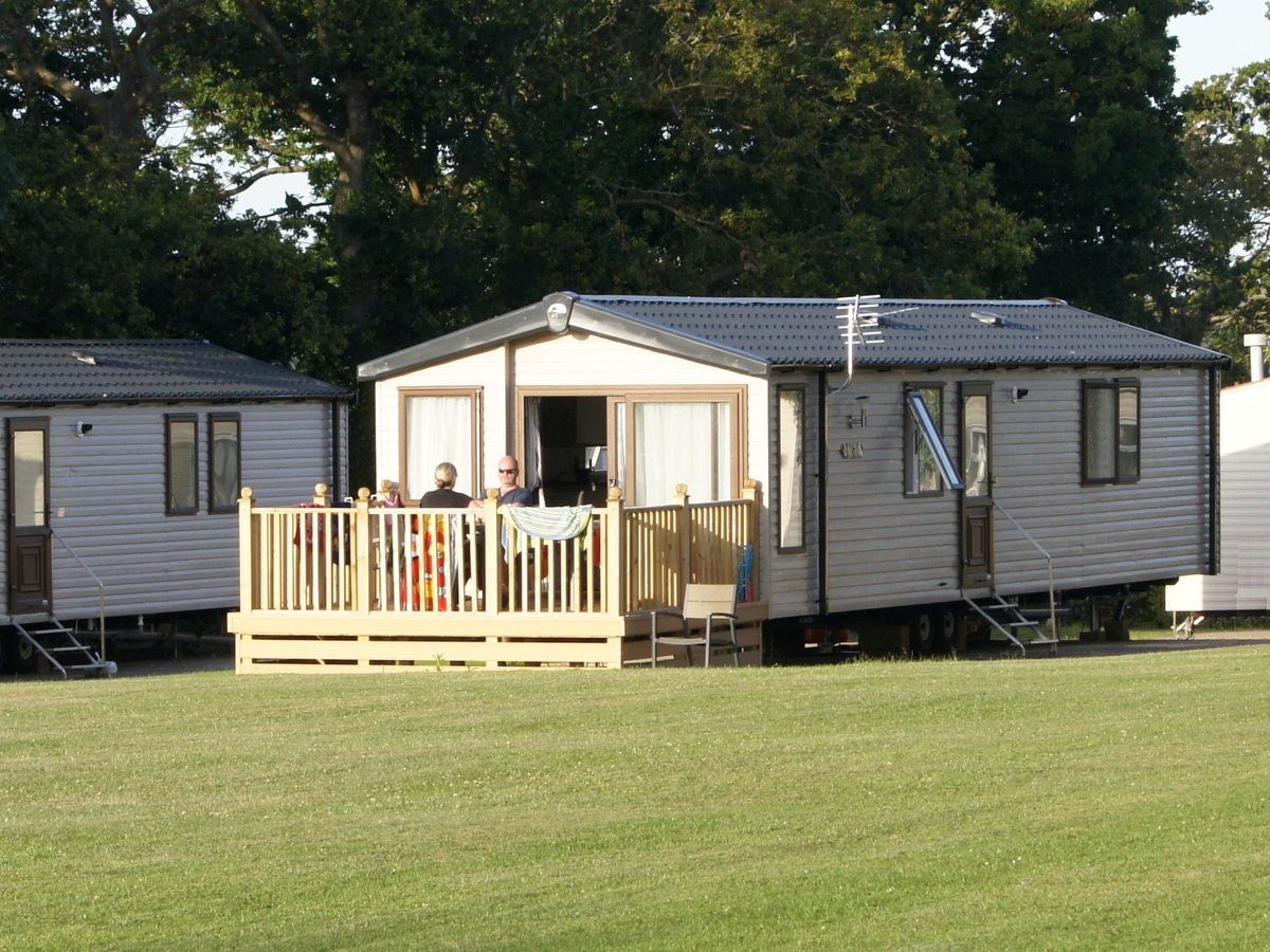 A cream static caravan with its patio doors open onto a timber decking where two people are sitting, on mown grass under tall trees.