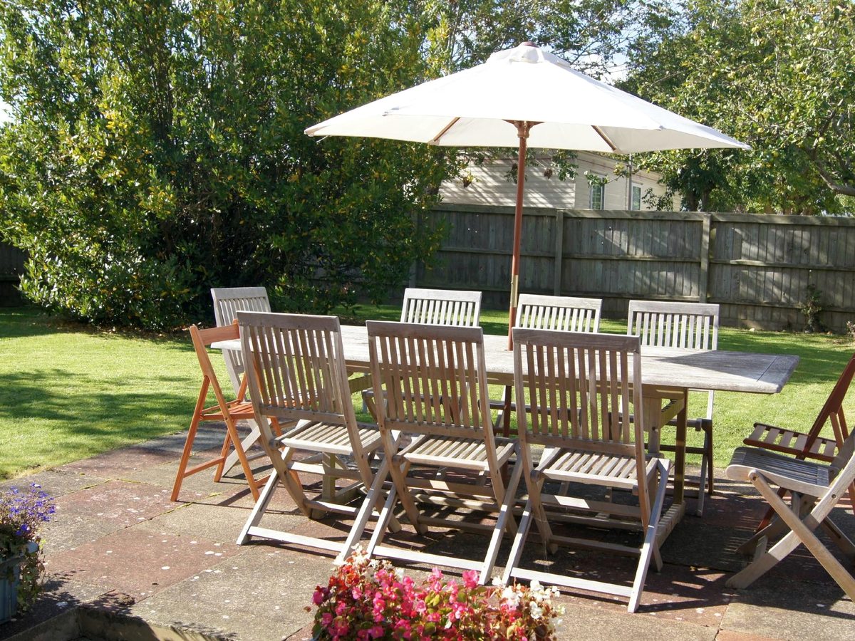 A wooden garden table with eight chairs under a cream parasol on a paved patio edged with flowerbeds and lawn.