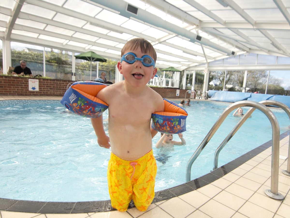 A young boy in orange armbands and goggles standing in the shallow end of an indoor pool under a glazed roof.