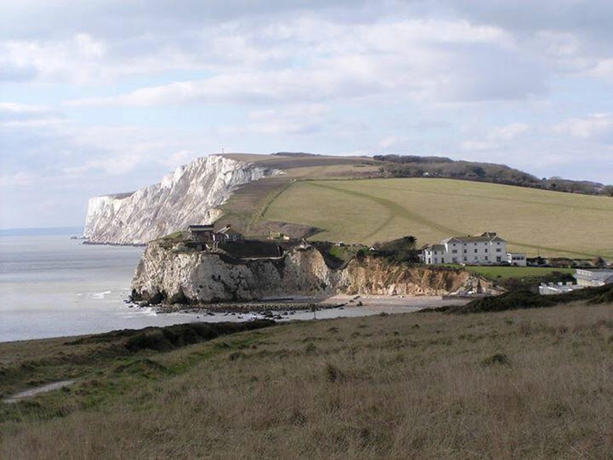 A grassy headland above a small bay, with a white chalk cliff rising behind a row of white buildings on the shore.