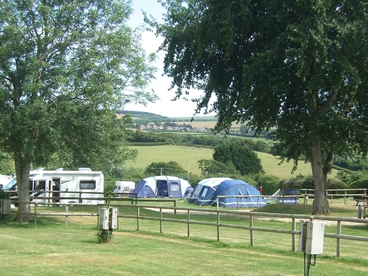 Blue family tents and a white motorhome on a grass touring field behind a post-and-rail fence, with rolling green farmland beyond.