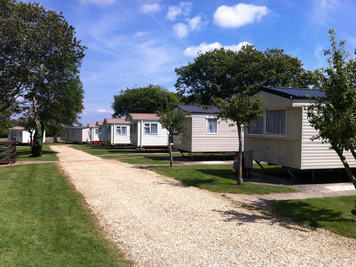 A gravel path running between rows of cream static caravans on mown grass, with mature trees and a blue summer sky.