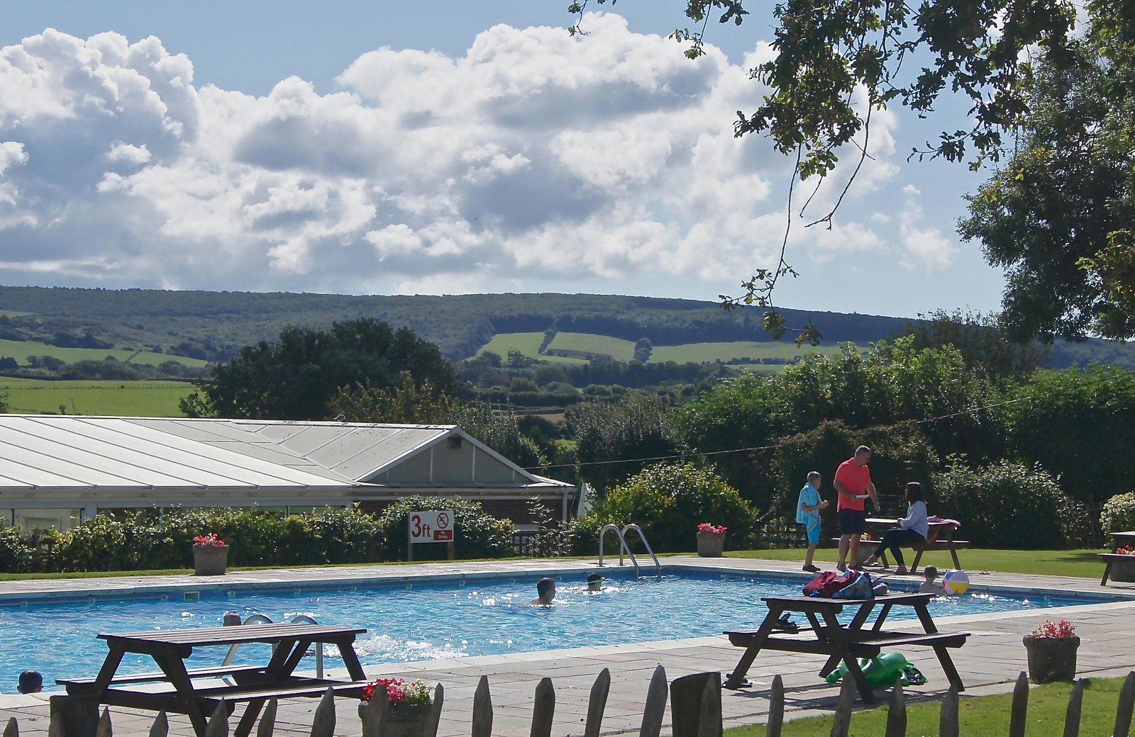 Children swimming in the outdoor pool at The Orchards Holiday Park, picnic tables on the paved surround, the glazed indoor pool building to one side and green downland behind.