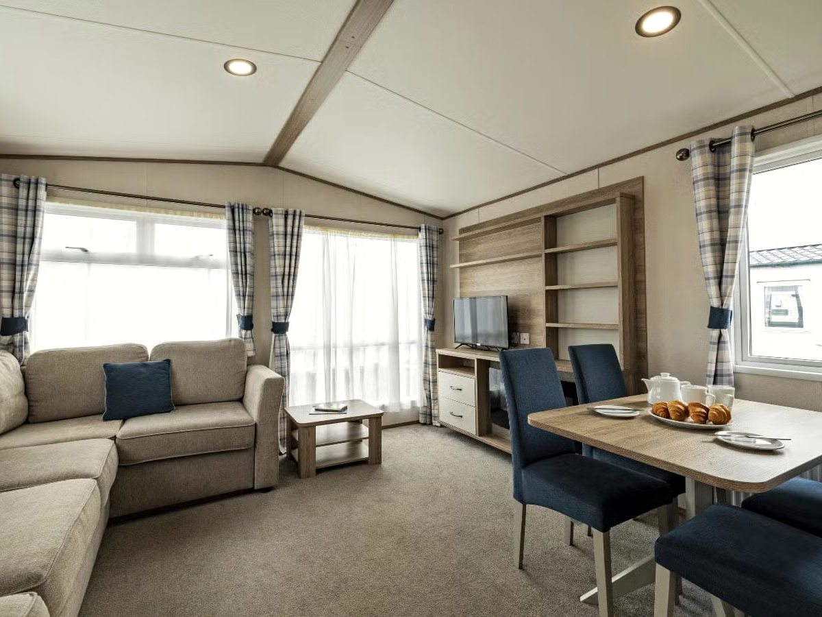 An open-plan caravan living room with a beige corner sofa, a low coffee table, a television on a wooden unit and a dining table with four blue chairs beside curtained windows.