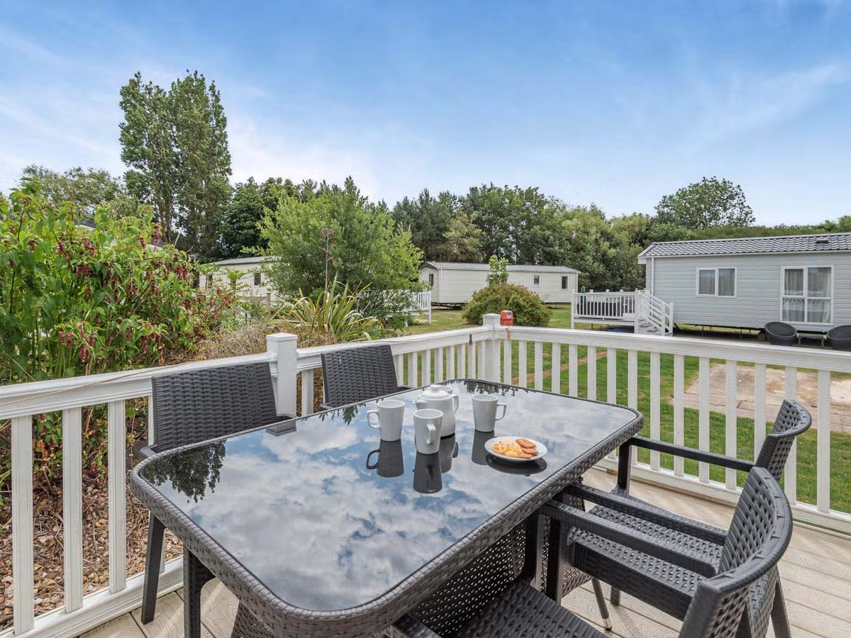 A raised timber deck with white railings, a glass-topped table and four woven chairs set with mugs and biscuits, with mown grass and other caravans beyond.