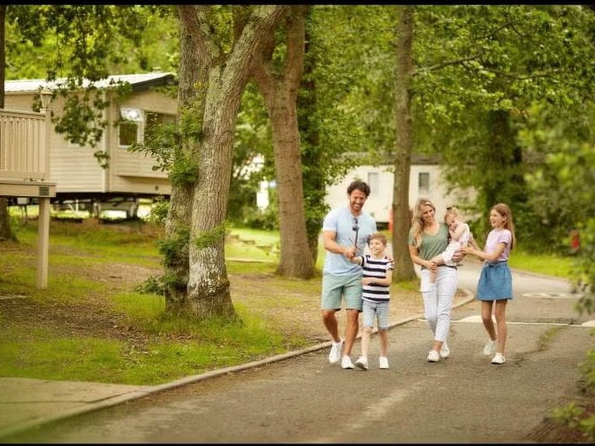 A family walking along a tarmac lane between static caravans under tall mature trees, with grass verges on either side.