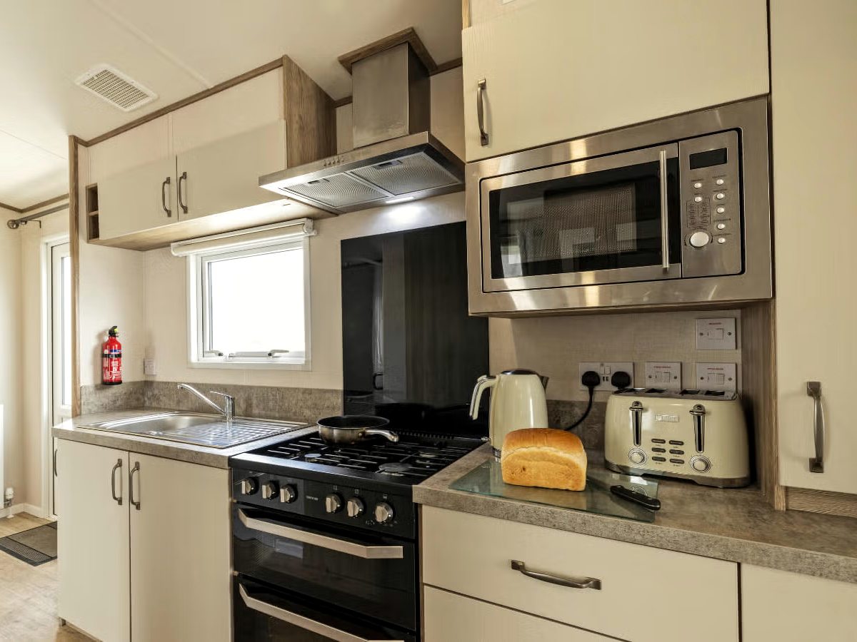 A fitted caravan kitchen with cream cupboards, a gas cooker under a stainless-steel extractor hood, a sink beneath a window, and a microwave, kettle and toaster on the worktop.