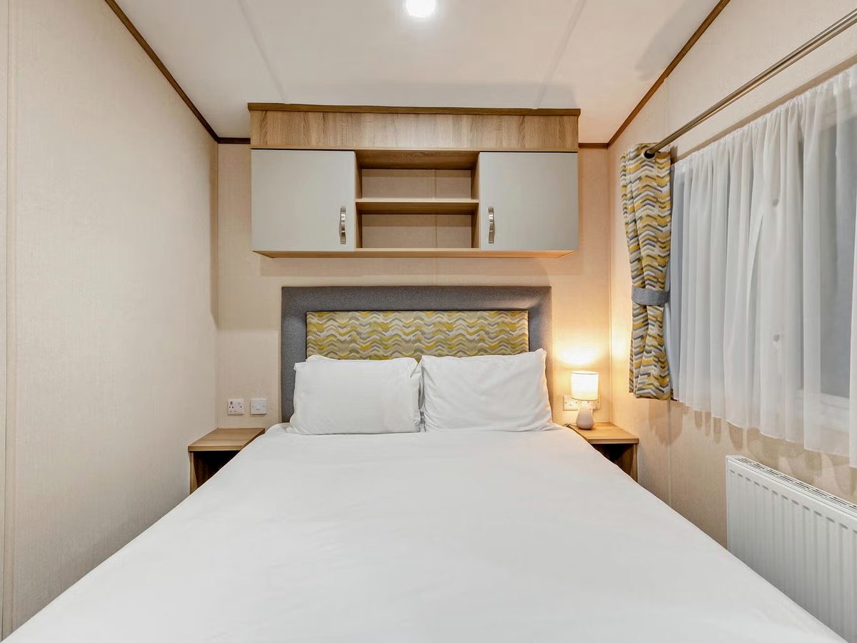 A double bed made up with white linen against a grey and yellow padded headboard, with overhead cupboards, narrow bedside tables and a curtained window alongside.