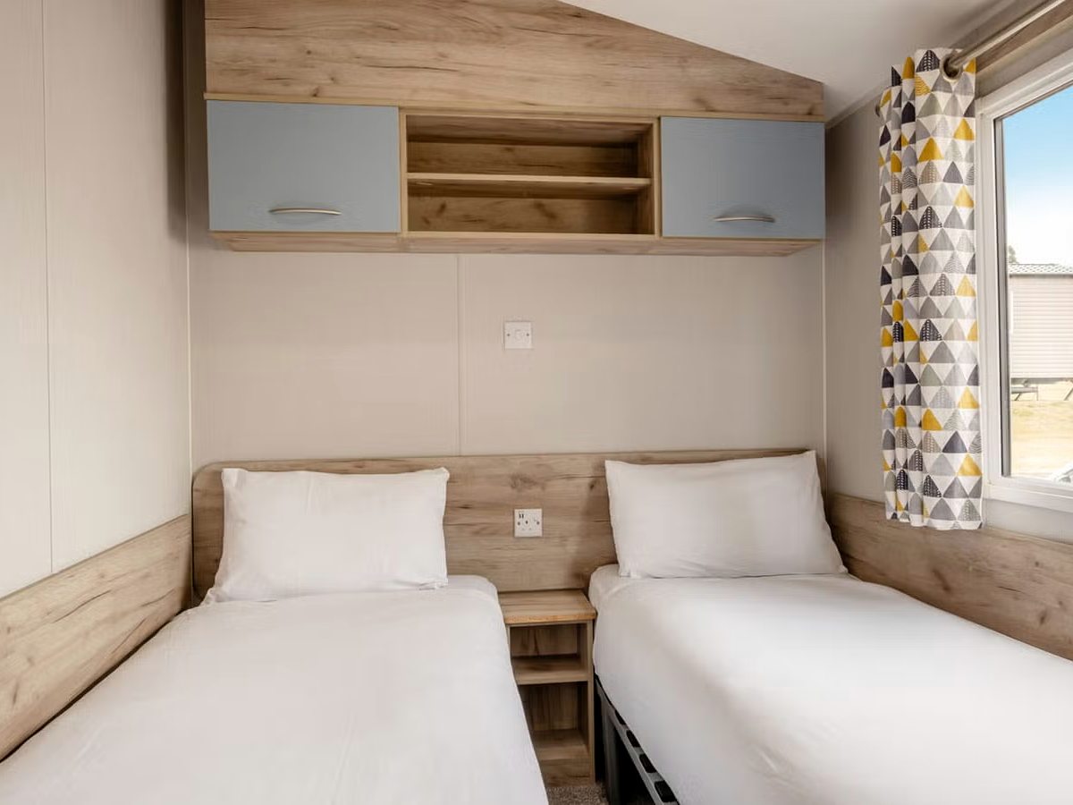 Two single beds with white bedding either side of a small bedside shelf, under a run of pale blue overhead cupboards and open shelving, beside a curtained window.