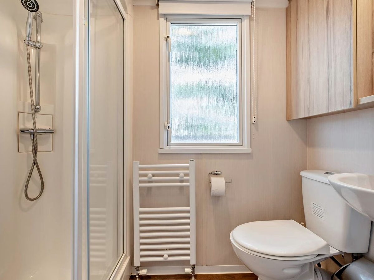 A caravan shower room with a glass shower enclosure and handheld shower, a white heated towel rail, a toilet and a washbasin under a frosted window.
