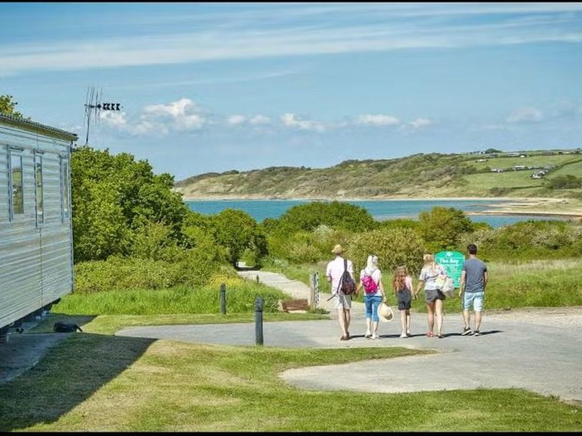 Five people walking down a tarmac path past a static caravan and a green signpost, with the bay and a green headland ahead of them.