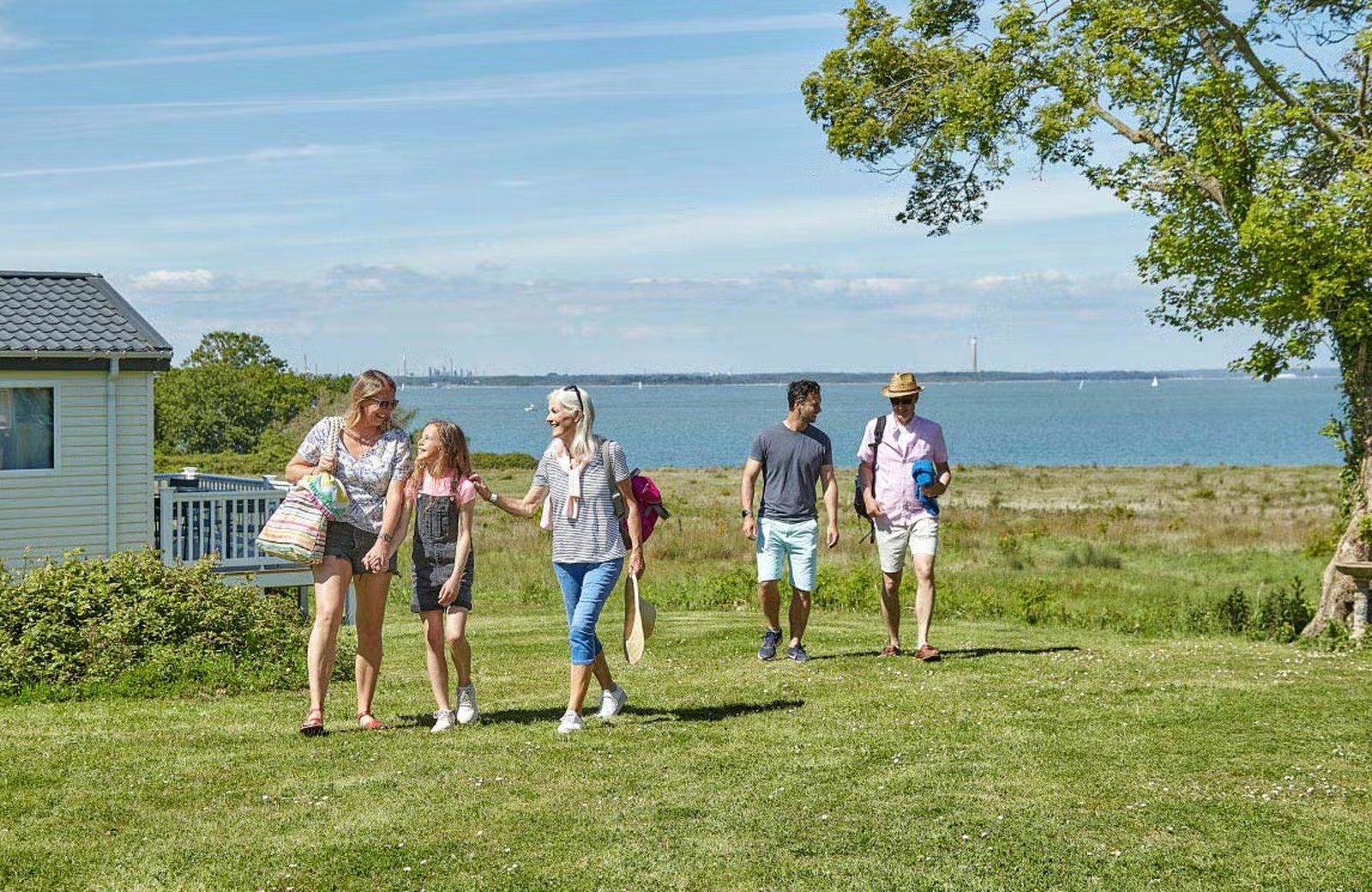 Families walking across mown grass at the bottom of Thorness Bay Holiday Park, with a static caravan and its decking to one side and the Solent and the Hampshire coast beyond.