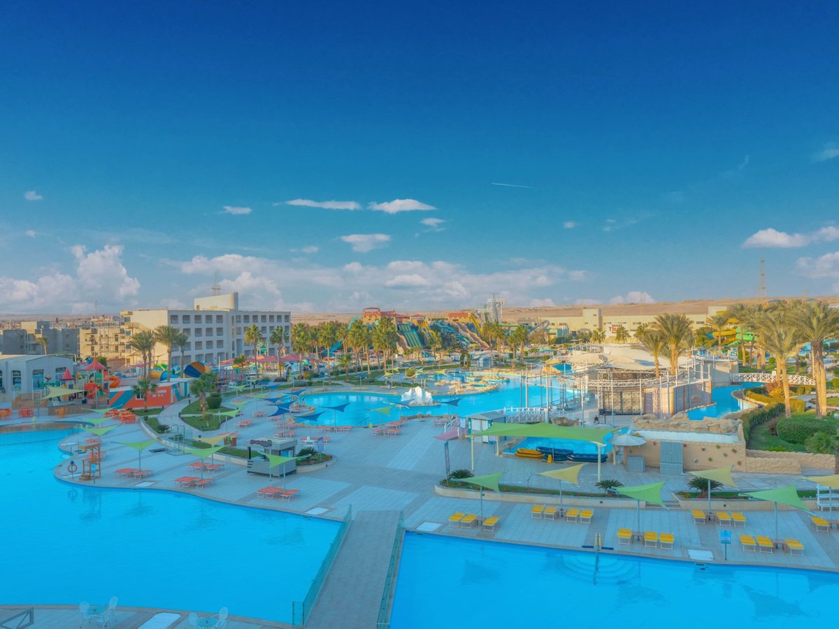An elevated view over a broad turquoise pool with yellow and orange loungers and green shade sails, and the aqua park's slide towers and children's area behind.