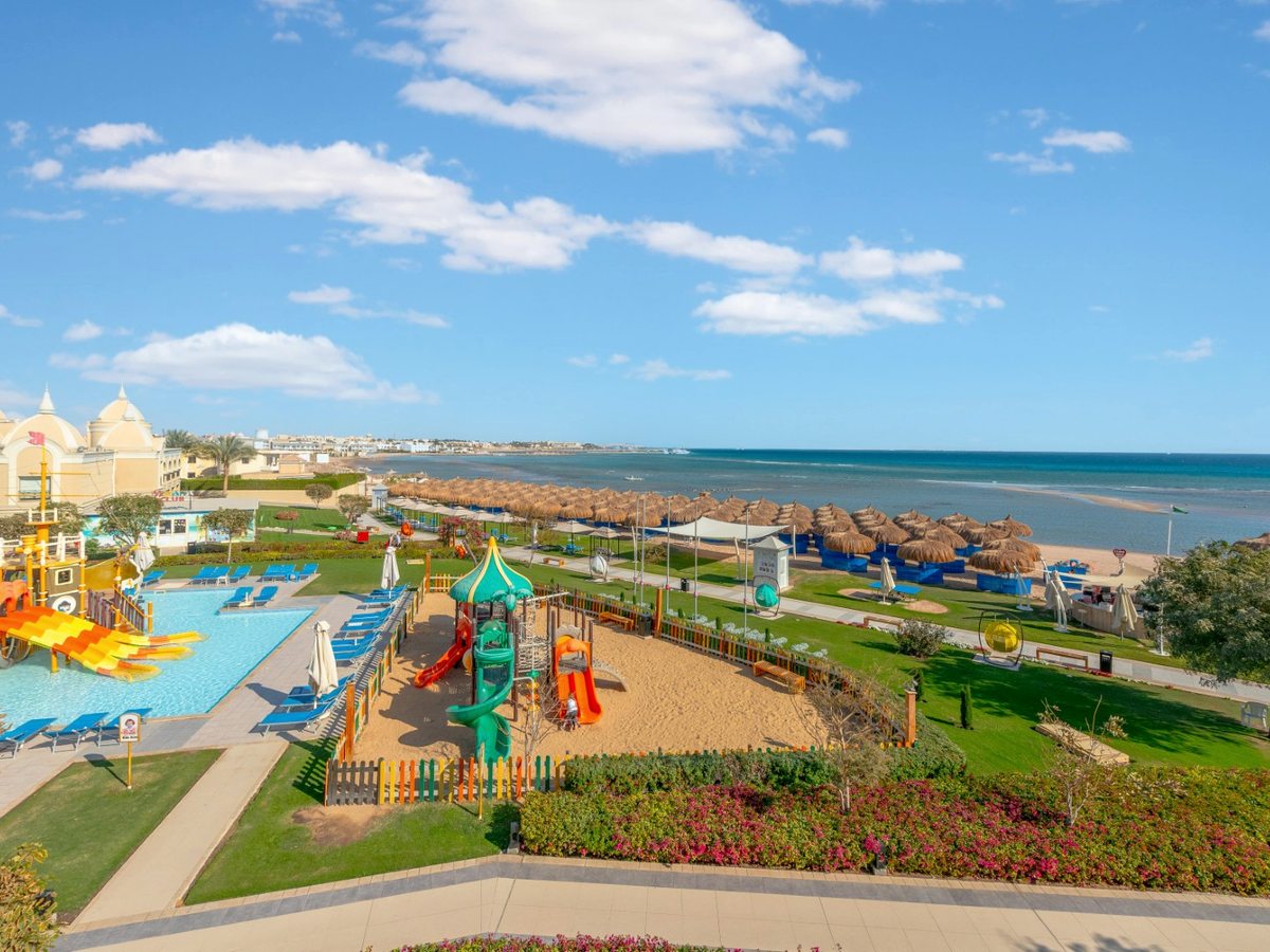 An elevated view of a fenced children's playground on sand next to a small pool with slides, with rows of thatched parasols along the beach and shallow turquoise sea beyond.