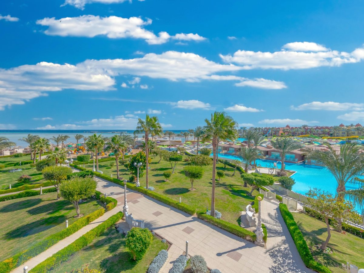 Landscaped lawns, clipped hedges, palms and a paved path in bright sun, with a pool and low bungalow rooms to one side and the sea in the distance.