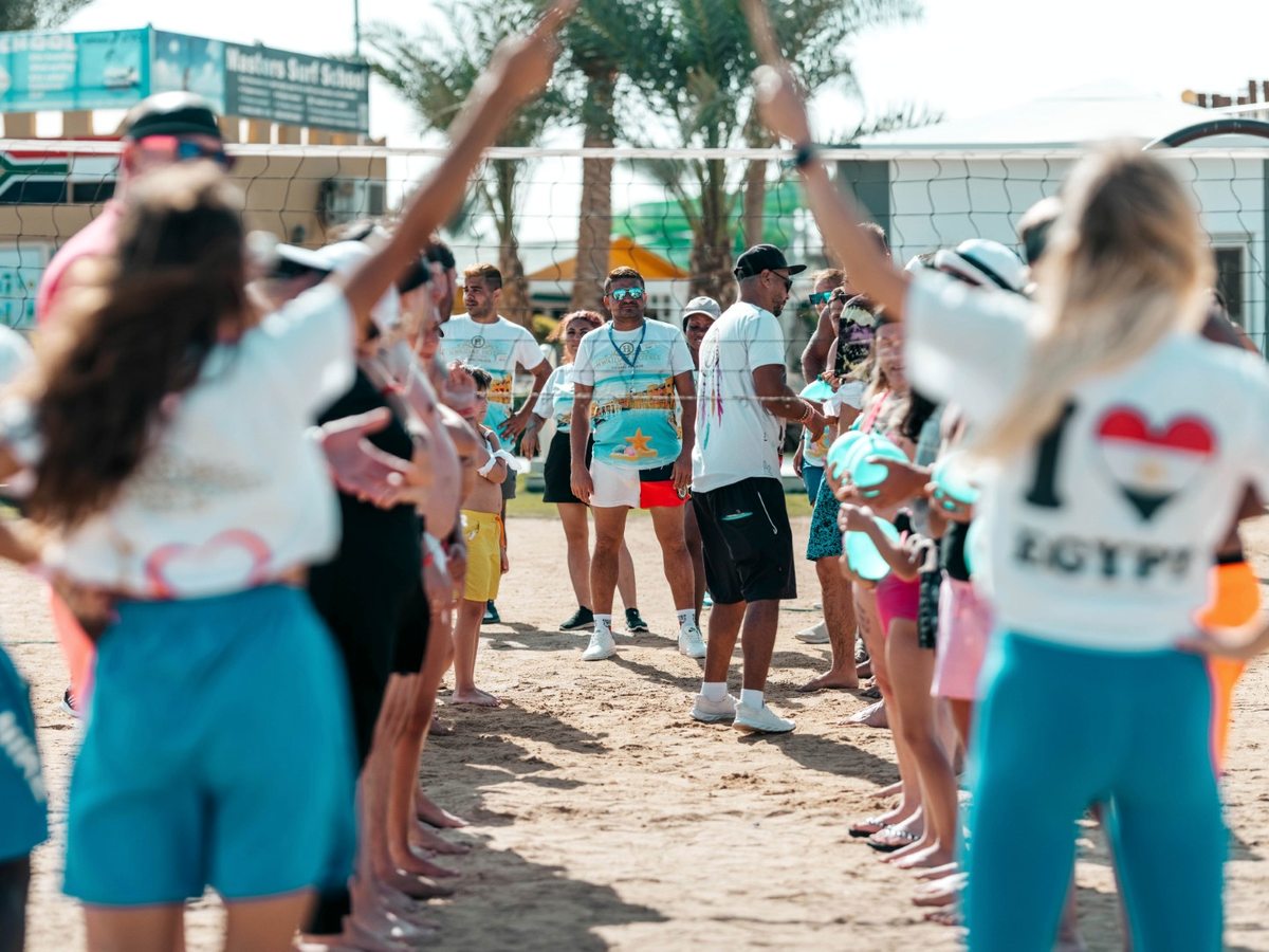 Holidaymakers standing in a circle on the sand around two animation staff by a volleyball net, arms raised, in bright sun.