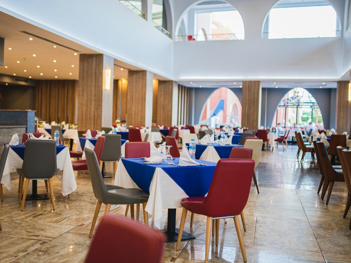 A large dining room with blue and white linen tables, red and grey chairs, a marble floor and tall arched windows, with a buffet counter along one side.
