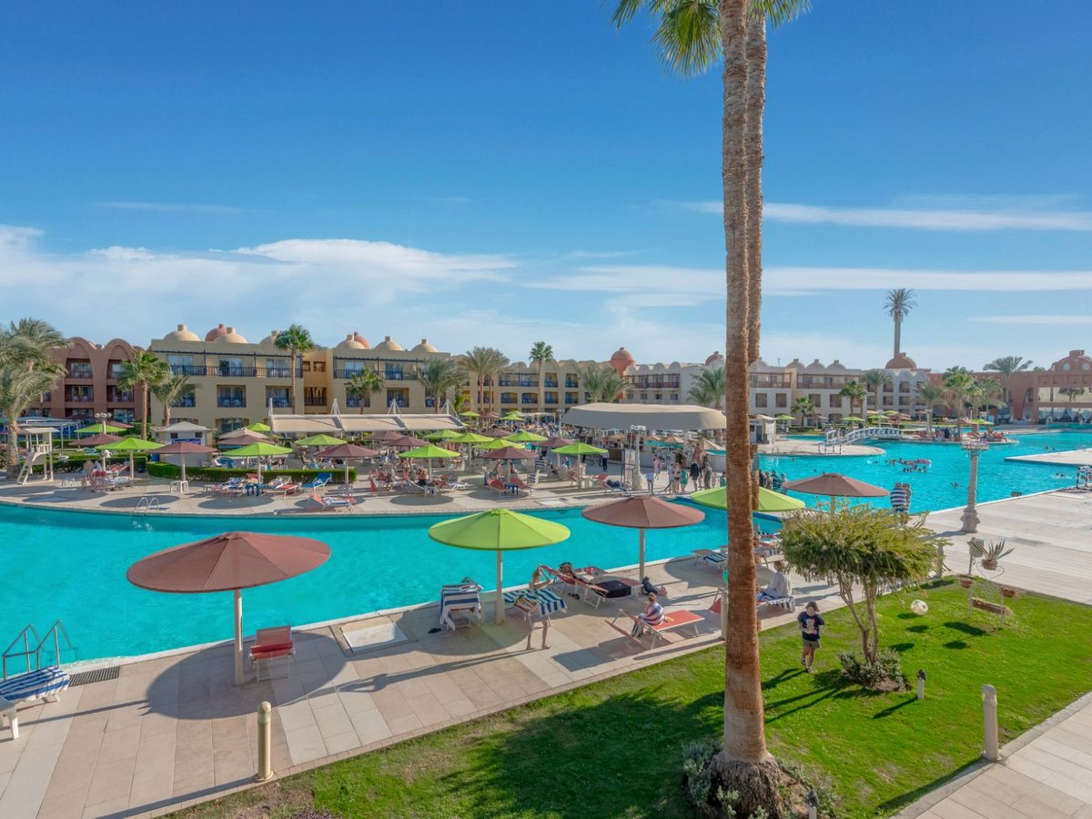 A pool edged by lawns and palms, with rows of sun loungers and open parasols along the paved terrace and guest buildings behind.