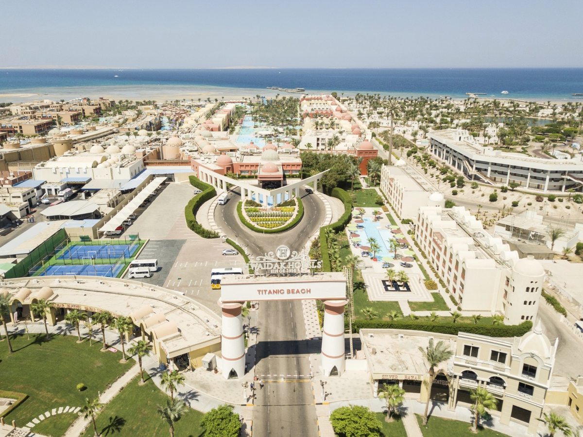 An aerial view of the resort's arched entrance gate lettered Titanic Beach, with the access road, tennis courts, low sand-coloured buildings and the Red Sea beyond.