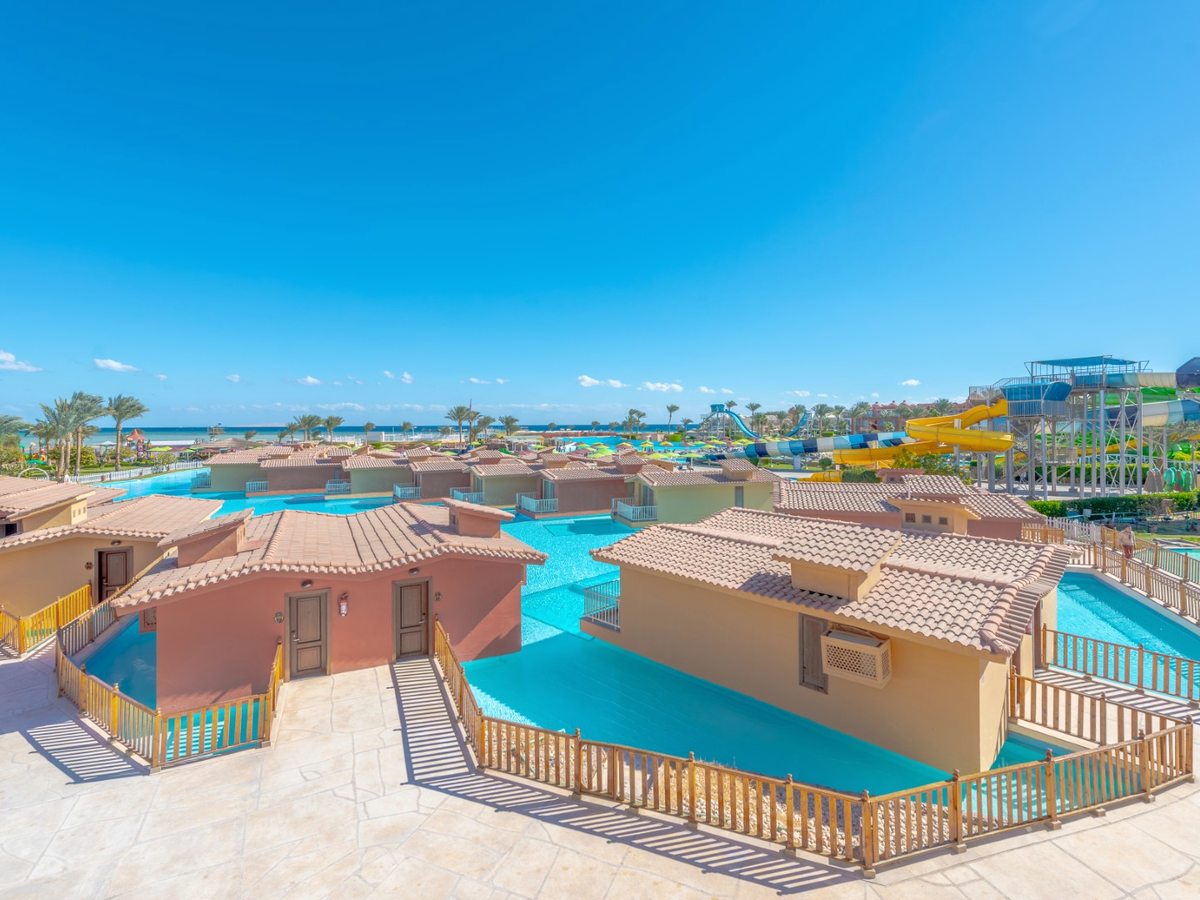 Rows of tile-roofed single-storey rooms with railed terraces at the edge of a shallow turquoise pool, the aqua park slides and the sea behind.