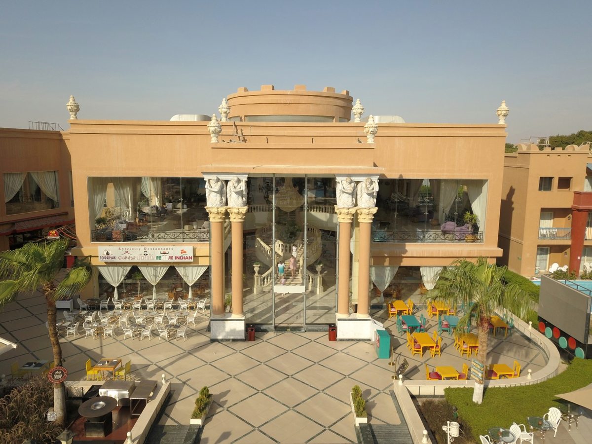 A sand-coloured restaurant building with tall columns and a glazed frontage, a sign listing the specialty restaurants, and a paved terrace of outdoor tables in front.